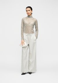 Μπλούζα με leopard print και μακριά μανίκια από διαφανές υλικό, συνδυασμένη με απαλές, ανοιχτογκρι παντελόνες με φαρδιά μπατζάκια. Κρατάει ένα μικρό λευκό τσαντάκι και φοράει μαύρες μυτερές ψηλοτάκουνες γόβες.