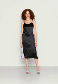 Vestido slip de cetim preto com decote em V, alças finas e corte até à panturrilha, combinado com uma bolsa lilás clara e sandálias.