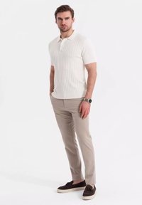 Polo s krátkým rukávem a žebrovanou texturou v krémové barvě, který je spárován s béžovými kalhotami na míru. Model má na sobě tmavě hnědé mokasíny a hodinky na levém zápěstí.