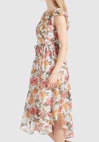 Eine Frau trägt ein ärmelloses, florales Midikleid mit Rüschensäumen und einem gebundenen Bund, während sie seitlich vor einem schlichten Hintergrund steht.