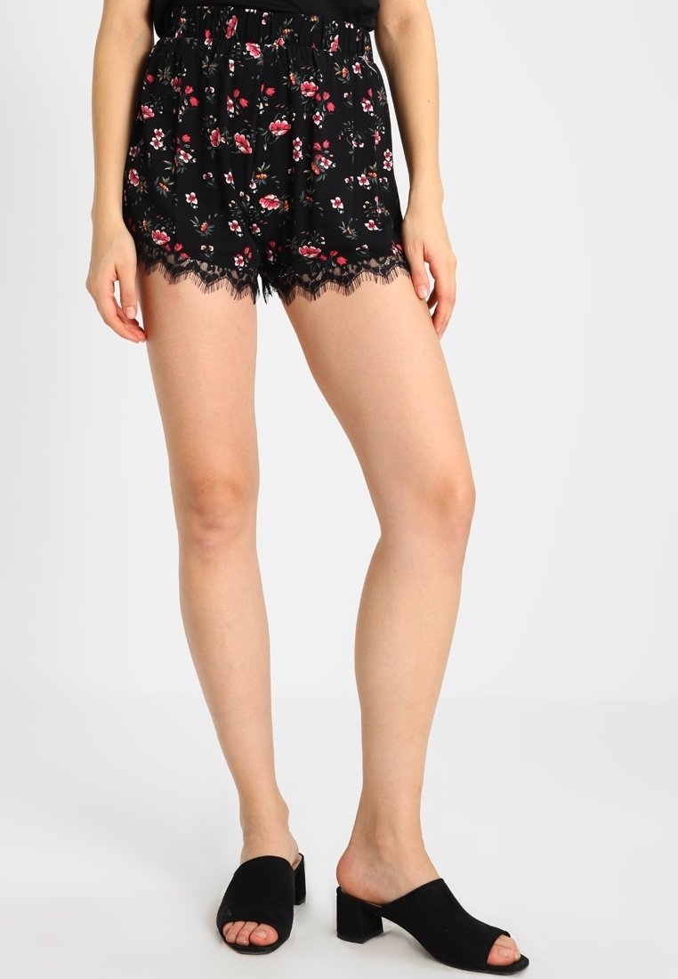 Schwarze Shorts mit Blumenmuster und gerafftem Bund, ausgestattet mit bunten Blumenmotiven und Spitzendetails. Kombiniert mit schwarzen offenen Clogs.