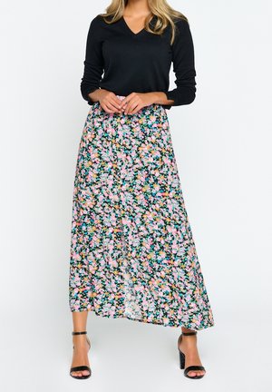 Femme portant un haut noir à manches longues, une jupe maxi fleurie avec des fleurs roses et bleues, et des talons noirs ouverts à bout avec bride à la cheville.