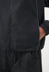 Gros plan d'une personne portant une veste noire zippée avec cordons et un pantalon noir, la main partiellement dans la poche de la veste.