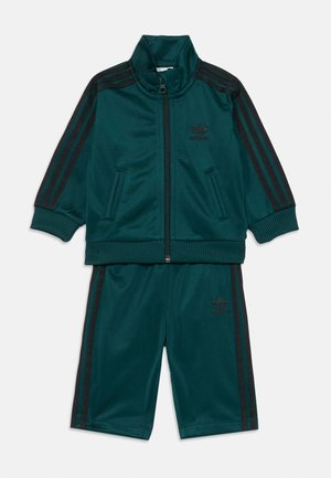 Ensemble de sport teal en tissu lisse. Comprend une veste zippée avec des rayures noires et un logo, ainsi que des shorts assortis avec des rayures latérales.