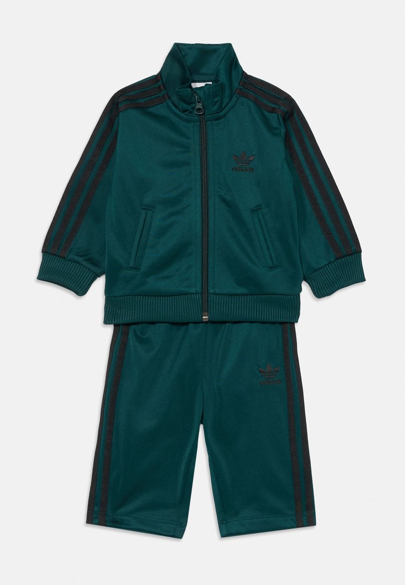 Ένα τυρκουάζ σετ φόρμας από άνετο ύφασμα. Περιλαμβάνει ζακέτα με φερμουάρ, όπως και μαύρες ρίγες και λογότυπο, και αντίστοιχα σορτς με πλευρικές ρίγες.