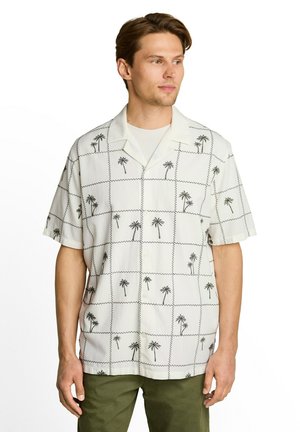 Man wearing a white short-sleeve button-up shirt with black palm tree and zigzag grid patterns, paired with olive green pants.