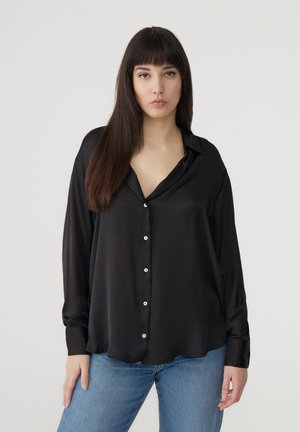 Blusa nera a maniche lunghe in tessuto liscio, con scollo a V, chiusura con bottoni bianchi e una vestibilità comoda. Abbinata a jeans in denim blu.