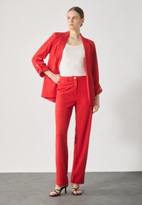 Rode blazer en bijpassende broek van gladde stof. De blazer heeft een enkele knop en opgerolde mouwen, gecombineerd met een witte top en zwarte sandalen.