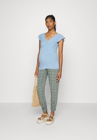 Top azul claro de mangas curtas com detalhes de babados, combinado com leggings verdes com padrão floral. Modelo segurando uma bolsa de palha, usando sandálias de plataforma.