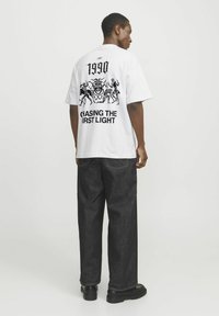 Hvid oversized t-shirt med sorte trykte grafik på ryggen, der viser "1990" og "CHASING THE FIRST LIGHT." Bæret med mørke denimbukser.