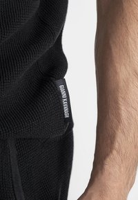 Indumento nero lavorato a maglia con struttura a coste, con un'etichetta di marca. Primo piano del materiale con un braccio abbronzato accanto.