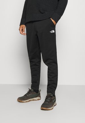 Osoba nosząca czarne spodnie dresowe The North Face, ciemne buty trekkingowe i ciemny sweter, stojąca na jednolitym jasnym tle.