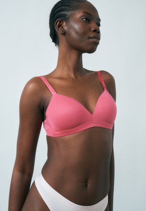 Femme portant un soutien-gorge rose et une culotte blanche, visible de la mi-poitrine jusqu'en haut, sur un fond uni gris clair.