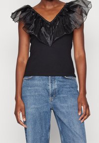 Kvinna i en svart ärmlös topp med genomskinlig volangkrage, matchad med högmidjade blå jeans, som står mot en neutral bakgrund.