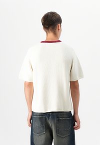Pulover cu mânecă scurtă din tricot crem, cu guler ribat maroniu. Dispune de o croială lejeră și un tiv drept, asortat cu blugi din denim închis.
