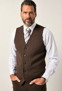 Braune Weste mit fünf Knöpfen, kombiniert mit einem weißen Hemd und einer grau gestreiften Krawatte. Die Hände in den Taschen, was einen strukturierten Stoff zeigt.