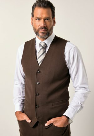 Chaleco marrón con cinco botones, combinado con una camisa blanca y una corbata a rayas grises. Manos en los bolsillos, mostrando una tela texturizada.