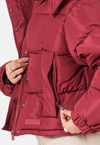 Burgundy puffjacka med en dragkedjeficka, tryckknappar, kviltad design och texturerat tyg med en logotyp på framsidan.