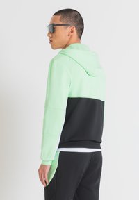 Felpa con cappuccio verde menta e nera dal texture liscia. Presenta un design a blocchi di colore e una tasca a marsupio. Indossata con occhiali da sole neri.
