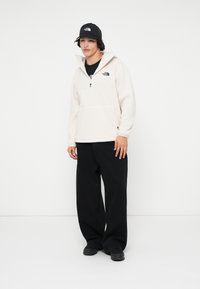 Giovane uomo che indossa un pullover bianco in pile e pantaloni neri, in piedi contro uno sfondo bianco semplice, con le mani rilassate ai lati.