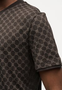 Καφέ πουκάμισο με κοντά μανίκια και επαναλαμβανόμενο κυκλικό γεωμετρικό σχέδιο που φοριέται από ένα άτομο σε λευκό φόντο.