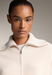 Pulover zip-up de un bej deschis cu un model texturat cu nervuri, guler înalt și detalii cu fermoar argintiu. Purtat peste un top alb.