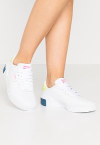 White sneakers with perforated texture, featuring a solid white sole, pink logo, and accents of light green and blue on the heel.
