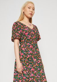 Robe florale à manches courtes noire, ornée de fleurs vibrantes roses, orange et vertes. Décolleté en V profond et coupe fluide.