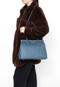 Blauwe leren handtas met een gestructureerde vorm, gouden hardware-details en een bovenhandvat, gedragen over een bruin bontjas.