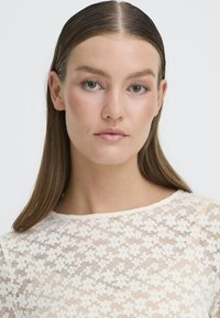 Blonder top med et blomstermønster i en blød kremfarve. Stoffet er gennemsigtigt med en let tekstur og en rund udskæring.
