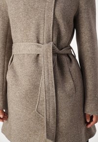 Manteau marron texturé avec ceinture à nouer à la taille, comprenant deux poches latérales et une coupe mi-longue. Le tissu présente un motif chiné subtil.