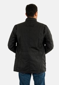 Zwarte gewatteerde jas met een hoge kraag, rugnaaddetails en een horizontale tab aan de nek. Draagt over blauwe jeans.