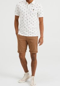 Bílé polo tričko s mnohobarevnými vzory, má límeček a krátké rukávy. V kombinaci s hnědými nakrčenými šortkami a bílými teniskami.