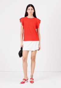 Haut rouge perforé à manches courtes avec des accents de volants, associé à une mini-jupe blanche. Le mannequin tient un sac à main noir et porte des chaussures à talons rouges.