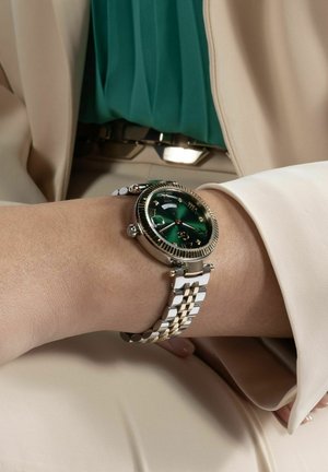 Polso con orologio in metallo argento e oro con quadrante verde e indicazione giorno-data, indossato sopra abito beige e camicetta turchese sullo sfondo.