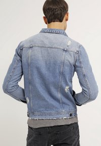 Man som bär en ljusblå slitna denimjacka över en grå skjorta, sedd bakifrån mot en enkel bakgrund.