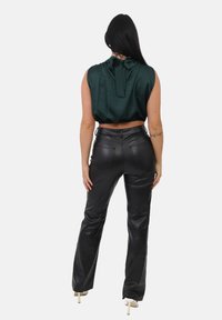 Kobieta z długimi ciemnymi włosami, ubrana w bezrękawnik w ciemnozielonym kolorze oraz wysokie czarne skórzane spodnie, stojąca tyłem do kamery.