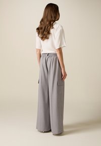 Mujer con cabello largo y ondulado, vistiendo una blusa blanca de manga corta y pantalones grises de pierna ancha y cintura alta con bolsillos laterales, de espaldas.