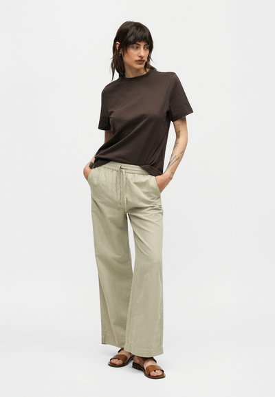 Γυναίκα με σκουρόμαυρα μαλλιά φοράει καφέ κοντομάνικο πουκάμισο, ανοιχτό μπεζ φαρδιά παντελόνια με κορδόνι και καφέ σανδάλια, στέκεται με τα χέρια στις τσέπες.