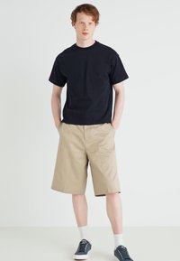 Black cotton t-shirt, beige knee-length shorts, white socks, and blue sneakers. Simple design and relaxed fit with no additional patterns.