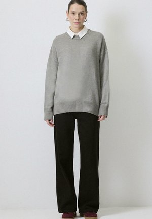 Kvinde stående iført en grå oversized sweater over en hvid skjorte med krave, sorte bukser med vide ben og bordeaux sko på en ensfarvet baggrund.