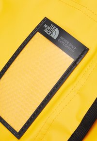 Yellow nylon fabric with a textured surface. Features a black rectangular label with a hexagonal pattern and white text: "THE NORTH FACE NEVER STOP EXPLORING."