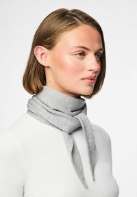 Grijze gebreide sjaal om de nek gewikkeld, met een gladde textuur en puntige uiteinden. Draagt bij een wit langemouwen shirt.