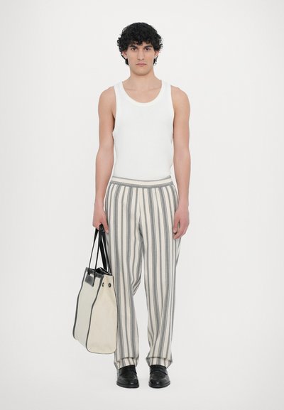 White tank top, striped baggy trousers with gray and beige stripes, carrying a black-handled tote bag, black footwear. Plain background.
