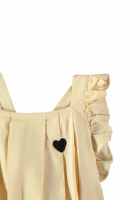 Gelbes Kleid mit gerüschten Schultern, plissiertem Vorderteil und einem kleinen schwarzen Herz-Patch auf der Brust. Weicher Stoff und zarte Textur.
