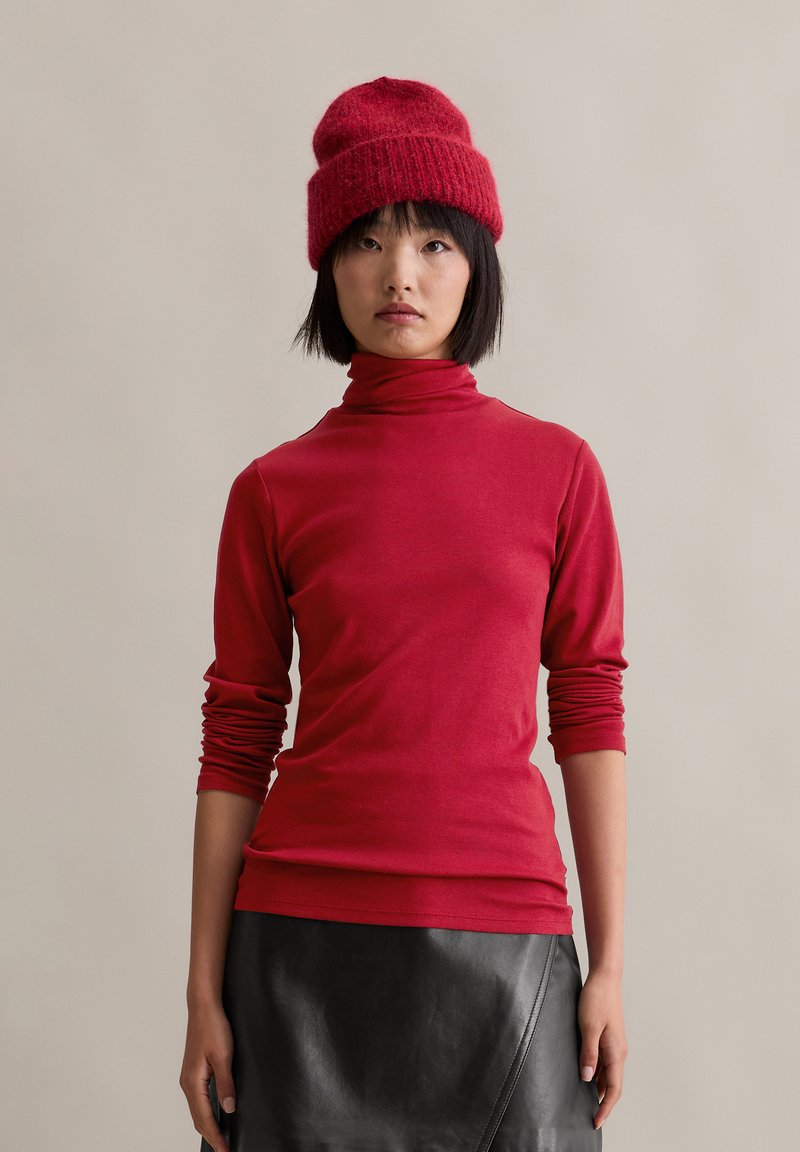 Red, long-sleeve turtleneck top with fitted design and soft texture, paired with a black leather skirt. Chunky red knit beanie on head.
