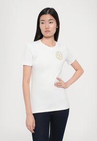 Camisa básica de manga curta branca com um decote redondo e um logótipo circular dourado no lado esquerdo do peito. O tecido parece suave e elástico.