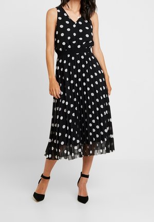 Femme portant une robe midi noire sans manches à pois blancs, un col en V, une jupe plissée et des talons noirs à bride cheville.