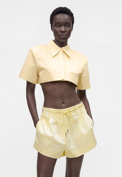 Femme à la peau foncée portant une chemise courte à manches courtes jaune pâle avec un col, et un short taille haute assorti avec poches.