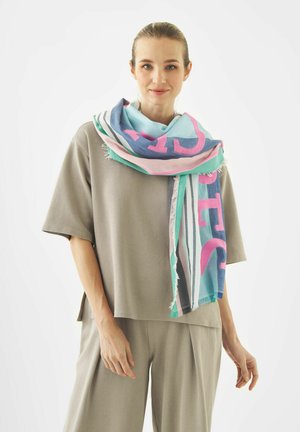 Junge Frau trägt beiges, weit geschnittenes Oberteil und Hose mit farbenfrohem, gestreiftem Schal um den Hals, steht vor einem schlichten Hintergrund.